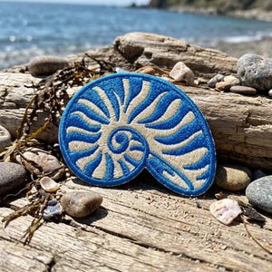 May include: A circular embroidered patch featuring a nautilus shell design. The embroidery consists of alternating curved lines in vibrant blue and off-white thread, creating a detailed spiral pattern that radiates from the center of the shell.