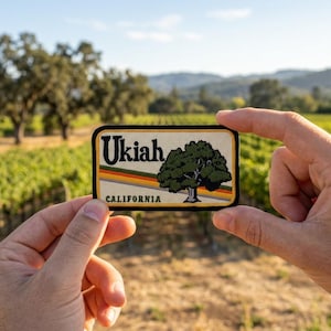 May include: A rectangular patch featuring the word "Ukiah" in black letters above a green tree illustration. Below the tree are horizontal stripes of orange, yellow, and brown. The word "CALIFORNIA" is printed below the stripes.