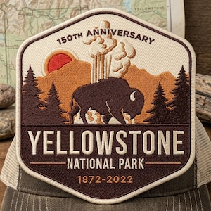 May include: A hexagonal embroidered patch featuring a brown bison in front of a geyser, orange sun, and pine trees. The text reads 150TH ANNIVERSARY, YELLOWSTONE NATIONAL PARK, and 1872-2022. The patch has a cream background with dark brown borders.
