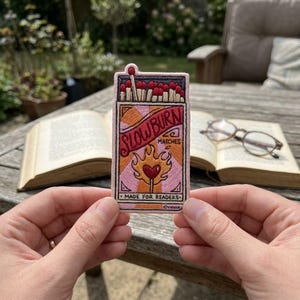 May include: Embroidered patch featuring a matchbox design with the text "SLOWBURN MATCHES" in a banner. The patch includes a heart-shaped flame and the words "MADE FOR READERS". The patch is held in front of an open book.