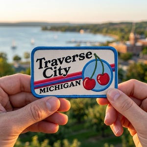 May include: A rectangular patch with a blue border features the text "Traverse City MICHIGAN" in black. A circular design with two red cherries and a blue stem is on the right. Red and blue stripes are below the text. The patch is held up by two hands.