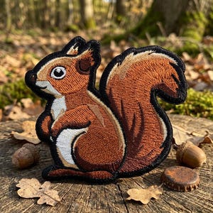 May include: Embroidered patch featuring a detailed squirrel design. The patch showcases a brown squirrel with a fluffy tail, white belly, and black outline. The patch is set against a natural background with acorns and leaves.