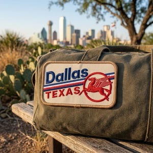 May include: An olive green canvas duffel bag featuring a rectangular embroidered patch. The patch displays the text Dallas in blue, Texas in red, and a red winged horse graphic inside a red circle, all set against a cream-colored background.