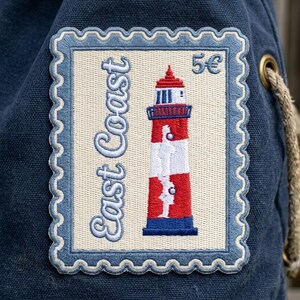 May include: A rectangular embroidered patch on a dark blue canvas fabric. The patch is shaped like a postage stamp with a cream background, featuring a red, white, and blue lighthouse, the text "East Coast" in blue, and the text "5€" in blue.
