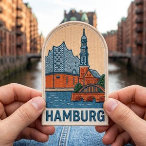 May include: An embroidered patch featuring a stylized depiction of the Hamburg skyline, including a tall church spire, brick buildings, and a bridge over water. The patch has a cream-colored background, a rounded top, and the word HAMBURG in blue.
