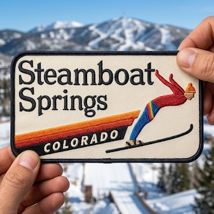 May include: Embroidered patch featuring the words "Steamboat Springs COLORADO" with a skier in mid-air. The patch has a cream background with a black border and a ski jump design in orange and red.