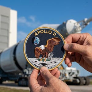 May include: A circular Apollo 11 patch featuring an eagle landing on the moon, with the Earth in the background. The patch has a navy blue background with gold trim and the words "Apollo 11" in gold lettering. The patch is held in front of a rocket.