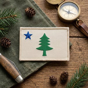 May include: A rectangular beige embroidered patch featuring a green pine tree centered on the fabric and a small blue five-pointed star positioned in the upper left corner. The patch has a stitched border and a clean, flat rectangular shape.