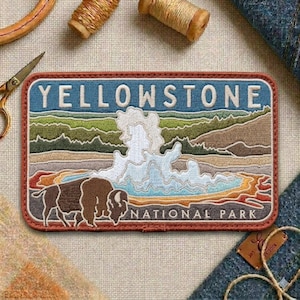 May include: Embroidered patch featuring a bison and a geyser in Yellowstone National Park. The patch has a blue background with the word "YELLOWSTONE" in white letters. The design includes a brown border and the words "NATIONAL PARK".