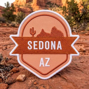 May include: A circular embroidered patch with a white border featuring a peach-colored background, a dark orange banner across the center with the white text SEDONA, and the white text AZ below. The design includes a saguaro cactus and rock formations.