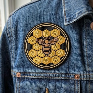 May include: Round embroidered patch featuring a honeybee design. The patch has a black border, a honeycomb pattern in gold, and a detailed bee in shades of brown and gold. The patch is affixed to a blue denim jacket.