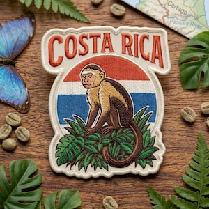 May include: An embroidered patch featuring a capuchin monkey sitting in green foliage against a background of red, white, and blue horizontal stripes. The text COSTA RICA is stitched in orange letters at the top of the off-white bordered patch.