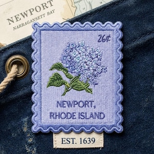 May include: A rectangular embroidered patch shaped like a postage stamp with a scalloped border. It features a light purple background, a blue hydrangea flower, and the text 26¢, NEWPORT, RHODE ISLAND, and EST. 1639 in dark purple thread.