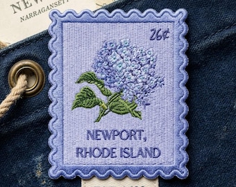 May include: A rectangular embroidered patch shaped like a postage stamp with a scalloped border. It features a light purple background, a blue hydrangea flower, and the text 26¢, NEWPORT, RHODE ISLAND, and EST. 1639 in dark purple thread.