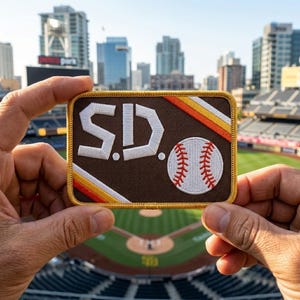 May include: A rectangular patch featuring the letters "S.D." in white, with a baseball graphic. The patch has a brown background with orange and yellow stripes. The patch is held in front of a baseball stadium.