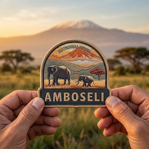 May include: Embroidered patch featuring a landscape with two elephants, a mountain, and the text "AMBOSELI NATIONAL PARK." The patch has a tan border and a black base with gold lettering. The background shows a grassy field and a mountain.