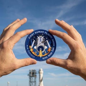 May include: A circular patch with the text "FIRST HUMAN FLIGHT" and "NEW SHEPARD" in a blue and white design. The patch features a rocket ship with flames and the names "BEZOS", "FUNK", and "DAEMEN".