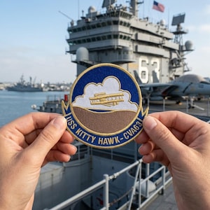 May include: A round patch with a blue border and the text "USS KITTY HAWK - CVA63". The patch features a cloud, a plane, and a brown landscape. The patch is held in front of a large aircraft carrier.