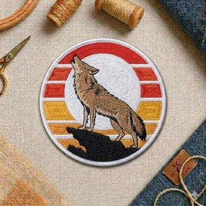May include: Embroidered patch featuring a howling wolf silhouette against a sunset backdrop. The patch is circular, with a white, red, and yellow color scheme. The wolf is brown, standing on a black rock. The patch is likely for clothing or accessories.