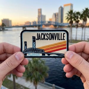 May include: A rectangular patch featuring the city name "JACKSONVILLE" in black block letters. The patch depicts a bridge with orange and yellow stripes, and the word "FLORIDA" is at the bottom. The patch is held between two hands.