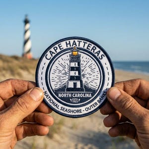 May include: A round patch featuring a lighthouse design with the text "Cape Hatteras" and "North Carolina". The patch is white and navy blue with a stitched border. The background shows a beach and a lighthouse.