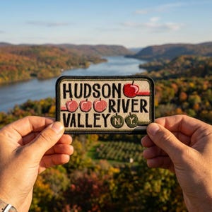 May include: A rectangular patch with the words "HUDSON RIVER VALLEY" in black letters, with red apple graphics. The patch also includes "N.Y." in green. The background shows a scenic view of a river and colorful trees.