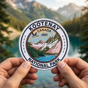 May include: A round embroidered patch featuring a scenic landscape of Kootenay National Park, Canada. The design includes mountains, a lake, and a flying bird, with the text "KOOTENAY NATIONAL PARK CANADA 1920" around the border. The patch is held up by two hands.