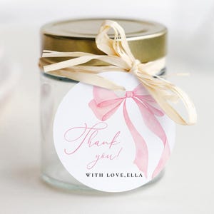 May include: A small glass jar with a gold lid, tied with raffia and a round tag. The tag features a watercolour pink bow and the words "Thank you!" and "WITH LOVE, ELLA" in pink script.