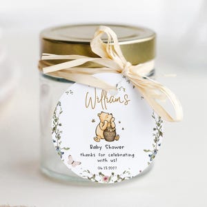 May include: A small glass jar with a gold lid, tied with a raffia bow. A round tag features a Winnie the Pooh illustration and the text "Williams Baby Shower thanks for celebrating with us! 06.13.2021". The tag is decorated with floral elements.