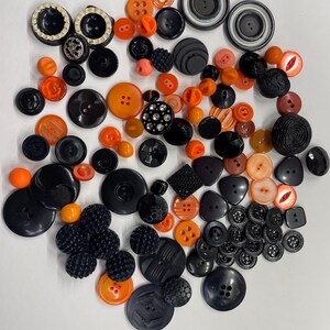 May include: A collection of assorted sewing buttons in various sizes, shapes, and textures, featuring a color palette of black, orange, and clear accents. The buttons include round, triangular, and square designs with two or four holes.