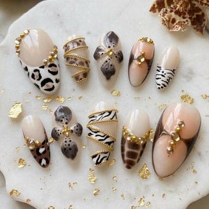 May include: A collection of ten almond-shaped artificial nails with various animal print designs. The nails feature leopard, zebra, and floral patterns, accented with gold embellishments. The color palette includes beige, brown, black, and gold.