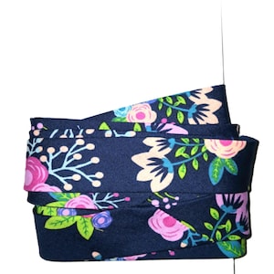 May include: A folded strip of dark navy blue fabric featuring a repeating floral pattern. The design includes stylized pink roses, light peach blossoms, bright green leaves, and small blue accents arranged across the smooth textile surface.