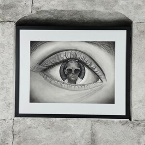 May include: Black and white framed art print featuring a detailed pencil drawing of a human eye. Inside the iris, a person wearing sunglasses is depicted. The artwork is mounted on a white mat and framed in a black frame, set against a textured gray wall.