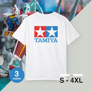 Puede incluir: Camiseta blanca con el logo de TAMIYA, dos estrellas rojas y azules y la palabra "TAMIYA" en azul. Disponible en tallas S-4XL y en 3 colores. Un modelo de robot es visible en el fondo.