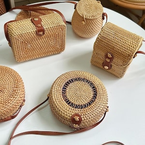 May include: A collection of woven rattan handbags in various shapes: a rectangular crossbody bag, a small round bag, a cylindrical bag, and a circular bag with a brown leather strap. The bags have brown leather accents.