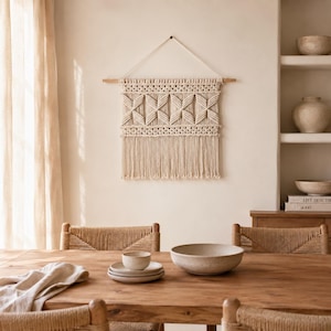 May include: A cream-colored macrame wall hanging featuring a geometric diamond pattern and long fringe, suspended from a horizontal wooden dowel by a white cord. The piece is crafted from thick cotton rope and displays intricate knotting techniques.