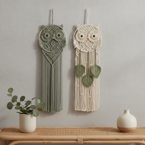 Op de afbeelding: Twee macramé uil wandkleden, één in saliegroen en één in crème, met ronde frames, geknoopte koordpatronen en houten kraaloogjes. De crème versie bevat drie bungelende macramé bladaccenten bevestigd aan de lange franjes.
