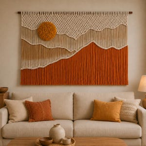 May include: A large rectangular macrame wall hanging featuring a layered landscape design. The piece uses cream, tan, and burnt orange cord, with a prominent circular textured mustard yellow sun element integrated into the upper left section.