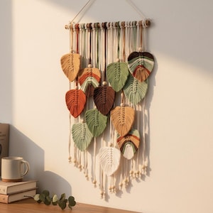 May include: A macrame wall hanging featuring leaf-shaped designs in various colors, including orange, green, and tan. The leaves are suspended from a wooden dowel with hanging cords and wooden beads, creating a decorative and textured wall art piece.