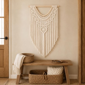 Könnte beinhalten: Ein cremefarbener Makramee-Wandbehang mit komplexen Knotenmustern, Holzperlen und einem Holzstab. Das Stück zeigt ein V-förmiges Design mit langen, fließenden Fransen, die spitz zulaufen und so ein strukturiertes Textilkunstwerk bilden.