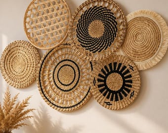 May include: A collection of six circular wall hangings made of woven natural fibers. The pieces feature various patterns including open lattice, concentric black and tan spirals, solid tan coils, and a sunburst design with black accents and fringe.