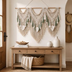 Op de afbeelding: Een grote macramé wandhanger met crèmekleurig koord, saliegroene accenten, houten kralen en drie bladvormige franje-elementen. Het kunstwerk hangt aan een horizontale houten roede en toont ingewikkelde knooptechnieken en diverse kwastjes.