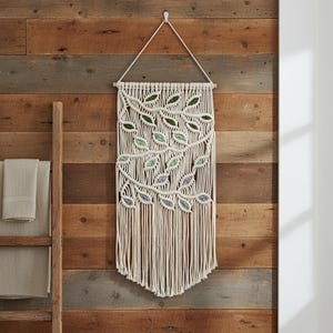 May include: A macrame wall hanging featuring off-white cotton cord knotted into a vine pattern with integrated leaves in shades of dark green, sage green, and dusty blue. The piece is suspended from a wooden dowel with a white hanging cord.