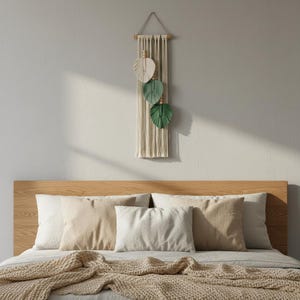 May include: A macrame wall hanging with three leaf accents in cream, green, and beige hangs above a bed with pillows and a blanket. The bed has a wooden headboard and is covered in neutral-toned bedding.