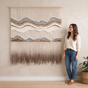 Peut inclure: Une grande tenture murale en macramé composée de cordes verticales couleur crème suspendues à une tige en bois. L'œuvre intègre des bandes horizontales tissées dans des tons bleu, beige et blanc, avec une frange dégradée brune en bas.