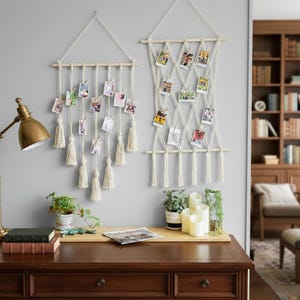 May include: Two macrame photo displays hanging on a gray wall. The displays are made of natural-colored rope and wooden dowels, with photos clipped to the strands. One has tassels. A desk with a lamp, plants, and candles is in the foreground.