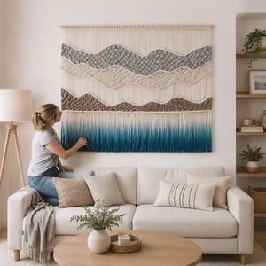 Op de afbeelding: Een grote rechthoekige macramé wandhanger met een reeks horizontale golvende patronen in grijstinten, crème en bruin, overgaand in een lange, dichte franje die verloopt van lichtblauw naar een diepe blauwgroene kleur aan de onderkant.