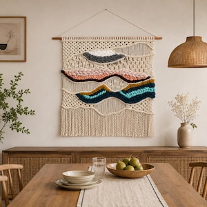 May include: A rectangular macrame wall hanging featuring a cream-colored cord base with woven horizontal sections in shades of white, gray, dusty pink, mustard yellow, and teal. The piece is suspended from a horizontal wooden rod with a white string.