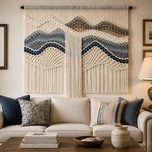 May include: A large rectangular macrame wall hanging featuring a series of undulating, layered waves in shades of cream, tan, blue, and dark navy. The piece is constructed from knotted cotton cord and hangs from a horizontal black rod.