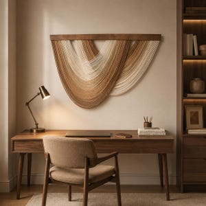 Puede incluir: Un tapiz de pared con una barra de madera horizontal y hebras de fibra en cascada en tonos crema, beige y marrón, dispuestas en curvas superpuestas en forma de U que crean un patrón texturizado y rítmico sobre la superficie de la pared.
